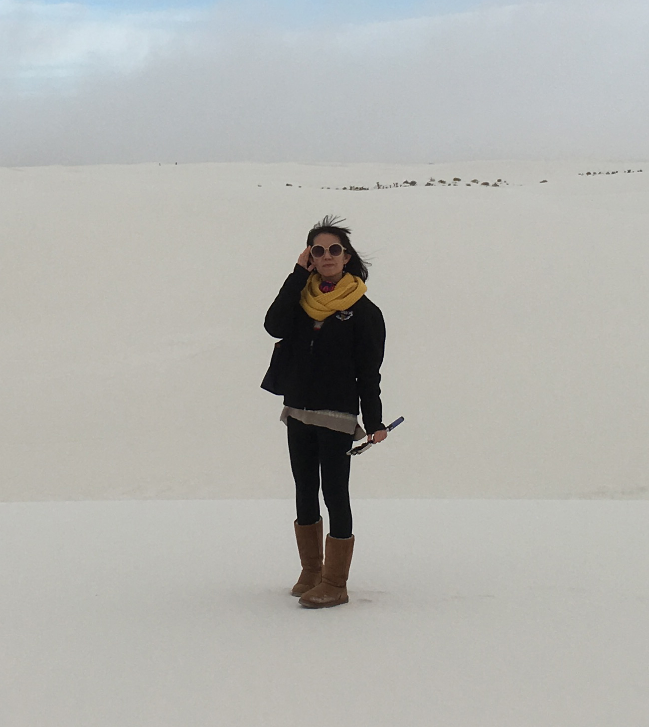 Crystal Yachin Lee stands on a sand dune, wearing a yellow scarf, round white glasses and brown boots. The sky is overcast and fog lingers in the background.